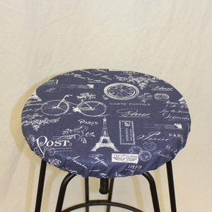 May include: A navy blue stool cover featuring a white Parisian design. The print includes the Eiffel Tower, bicycles, clocks, and the word "Paris". The cover is fitted over a black metal stool frame.
