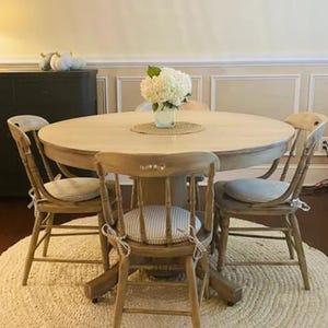 May include: A round wooden dining table with a light natural finish, accompanied by four matching spindle-back chairs. Each chair features a beige and white striped seat cushion tied with fabric ribbons. A white hydrangea bouquet sits in a glass vase.