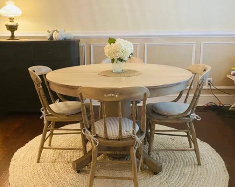 May include: A round wooden dining table with a light natural finish, accompanied by four matching spindle-back chairs. Each chair features a beige and white striped seat cushion tied with fabric ribbons. A white hydrangea bouquet sits in a glass vase.
