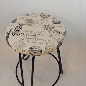 Pu&ograve; includere: Uno sgabello in metallo nero con una seduta rotonda rivestita in tessuto beige. Il tessuto presenta una stampa in bianco e nero di francobolli e scritte parigine vintage, tra cui il testo "2 AOUT 1873" e "PROVOT FRERES".