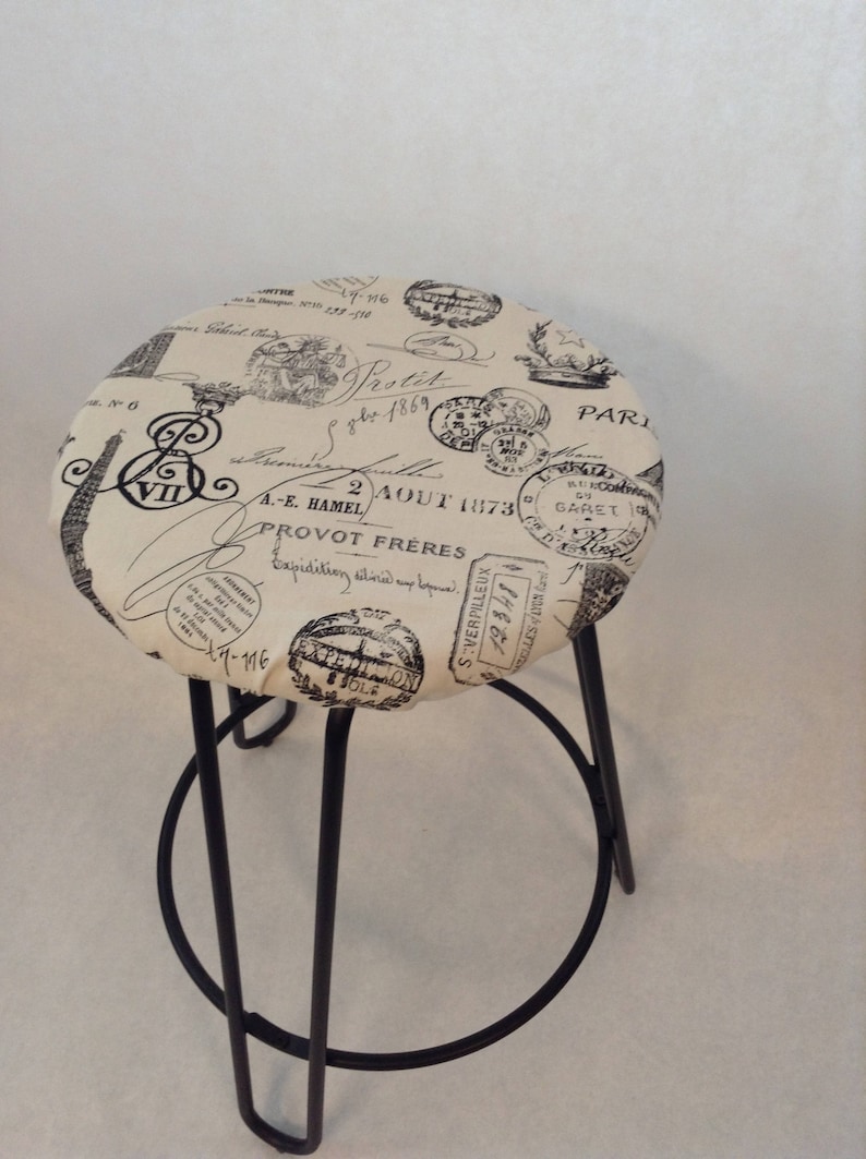 Pu&ograve; includere: Uno sgabello in metallo nero con una seduta rotonda rivestita in tessuto beige. Il tessuto presenta una stampa in bianco e nero di francobolli e scritte parigine vintage, tra cui il testo "2 AOUT 1873" e "PROVOT FRERES".