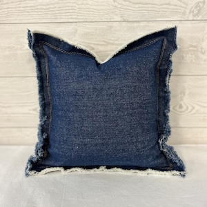 May include: A square denim pillow with a frayed edge. The pillow is a deep indigo blue with a lighter, frayed edge. The pillow is a square shape and appears to be made of a sturdy denim fabric. The pillow is a decorative item.