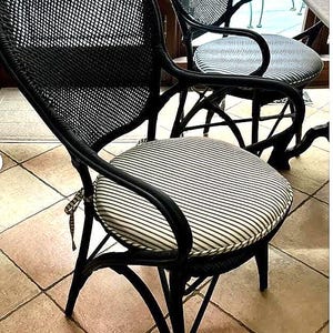 May include: A black woven armchair featuring a curved backrest and a round seat cushion with a fine black and white pinstripe pattern. The chair has integrated armrests and a tied fabric attachment on the side of the seat cushion for securing.