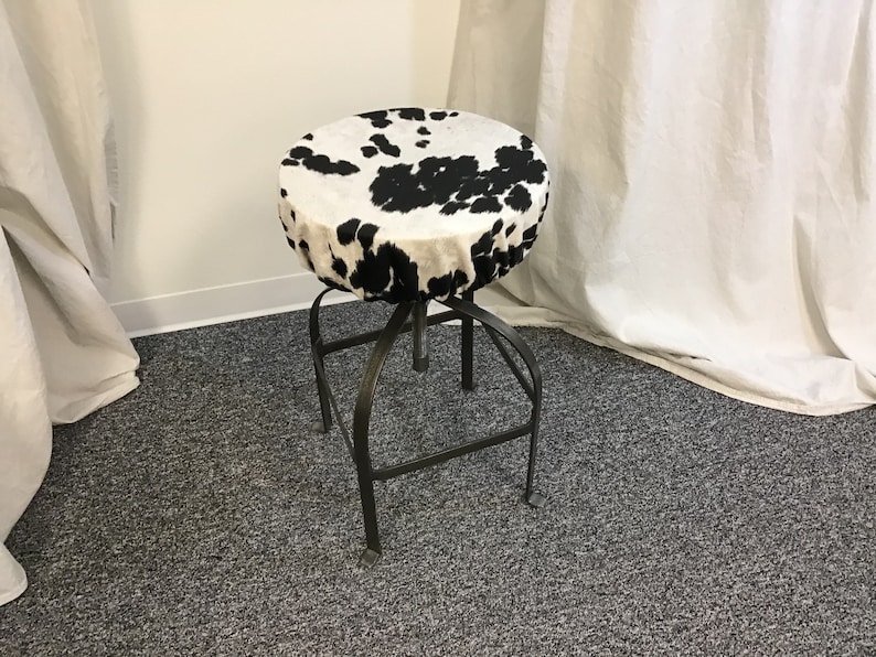 Pu&ograve; includere: Uno sgabello in pelle di mucca bianco e nero con base in metallo. Lo sgabello &egrave; su un tappeto grigio.