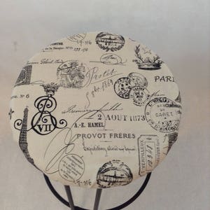 Pu&ograve; includere: Uno sgabello rotondo color crema con struttura in metallo nero. La seduta &egrave; rivestita in tessuto con un design parigino vintage con timbri e testo neri, tra cui "Protit" e "2 AOUT 1873".