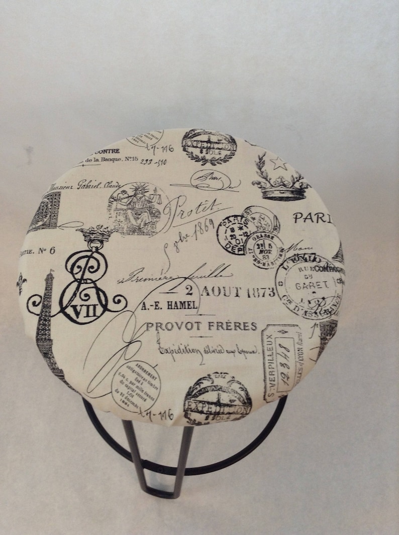 Pu&ograve; includere: Uno sgabello rotondo color crema con struttura in metallo nero. La seduta &egrave; rivestita in tessuto con un design parigino vintage con timbri e testo neri, tra cui "Protit" e "2 AOUT 1873".