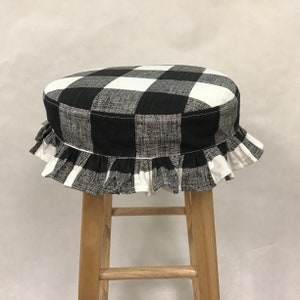 May include: A black and white checkered stool cover with a ruffled edge. The cover is placed on a wooden stool. The pattern is a classic buffalo check design, adding a touch of farmhouse style to the decor.