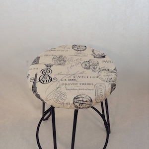 Pu&ograve; includere: Uno sgabello in metallo nero con una seduta rotonda rivestita in tessuto beige con una stampa in bianco e nero di francobolli e scritte parigine vintage. Il testo include "Protit gbe 1869", "2 AOUT 173", "A.-E. HAMEL", "PROVOT FRERES", "Expetition dilinde" e "17-176".