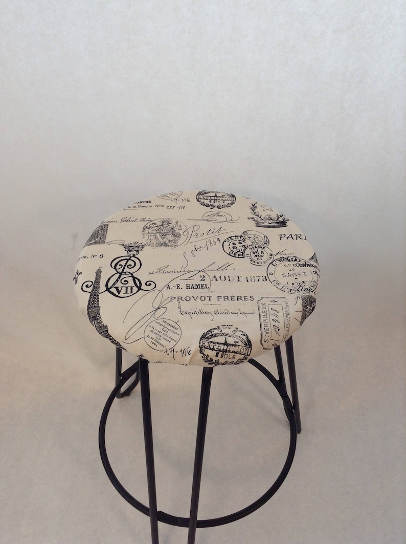 Pu&ograve; includere: Uno sgabello in metallo nero con una seduta rotonda rivestita in tessuto beige con una stampa in bianco e nero di francobolli e scritte parigine vintage. Il testo include "Protit gbe 1869", "2 AOUT 173", "A.-E. HAMEL", "PROVOT FRERES", "Expetition dilinde" e "17-176".
