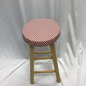 Könnte beinhalten: Ein Holzhocker mit einer runden Sitzfläche, die mit einem rot-weiß karierten Stoff bezogen ist. Der Hocker hat vier gerade Beine, die durch horizontale Verstrebungen verbunden sind und eine stabile Basis für das runde Polster bilden.