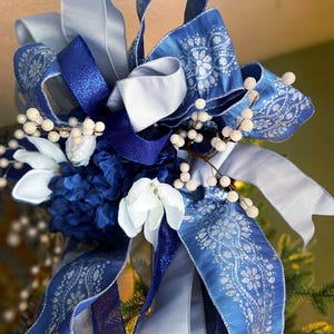 Puede incluir: Un lazo decorativo con cintas superpuestas en tonos azul real, azul claro y plata. El diseño incluye cintas estampadas con motivos florales plateados, cintas azules brillantes y cintas lisas de color azul claro, acentuadas con flores blancas.