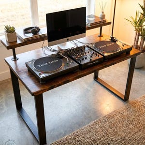 May include: A wooden DJ desk with black metal legs, featuring two Technics turntables, a Pioneer DJ mixer, and a computer monitor. A shelf above holds headphones and a small plant. The desk is in front of a window.