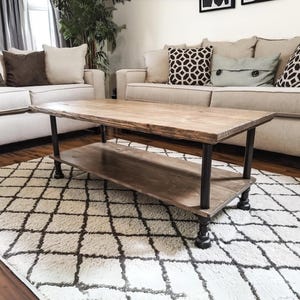 May include: A rustic wooden coffee table with a lower shelf, featuring black metal pipe legs. The table is placed on a white and brown patterned rug in a living room setting.