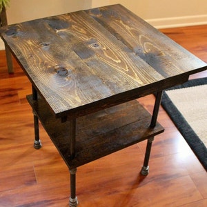May include: A dark brown wooden end table with a lower shelf and black metal pipe legs. The table is sitting on a hardwood floor.