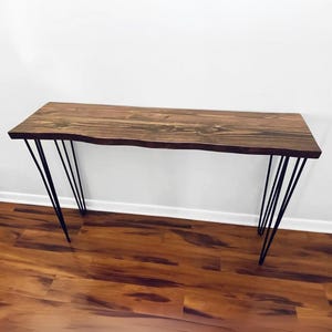May include: A wooden console table with a live edge top and black hairpin legs. The tabletop is stained a dark brown, showcasing the natural wood grain. The table is approximately 60 inches long.