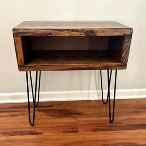 May include: A dark brown wooden end table with a shelf and hairpin legs. The table has a rustic, reclaimed wood look.