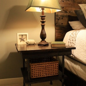 May include: A wooden end table with a dark brown finish and black metal legs. The table has a top shelf and a lower shelf that holds a woven basket. A lamp with a green lampshade sits on the top shelf, and a book sits on the table next to the lamp.