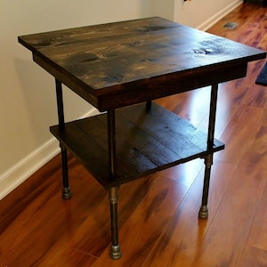 May include: A dark brown wooden side table with two shelves and black metal pipe legs. The table is standing on a hardwood floor.