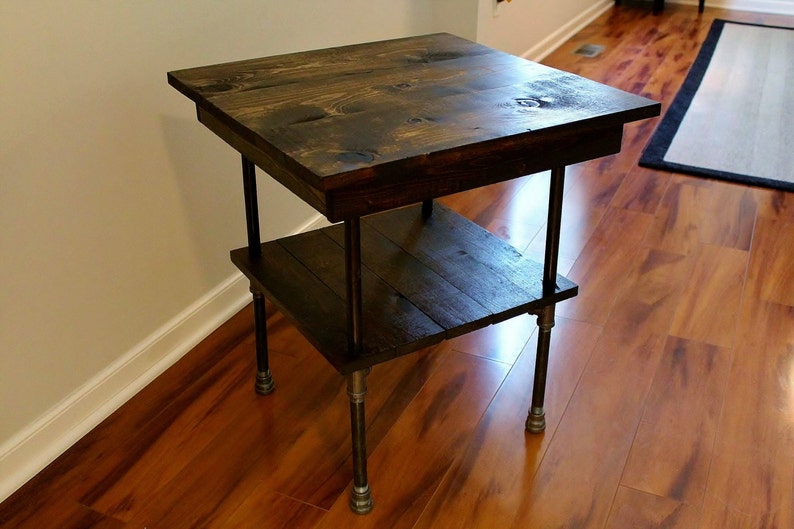 May include: A dark brown wooden side table with two shelves and black metal pipe legs. The table is standing on a hardwood floor.