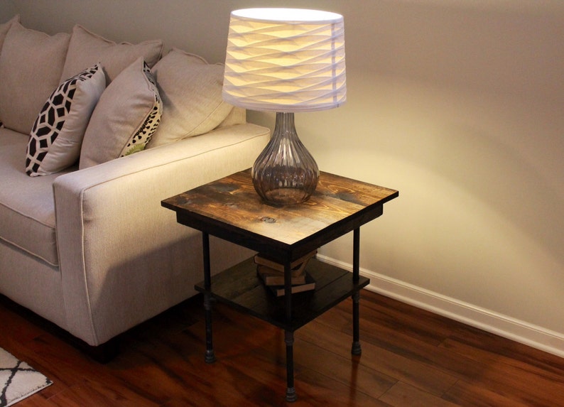 May include: A rustic wooden end table with a black metal frame and a white lamp with a pleated shade. The table has a shelf for storage.