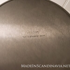 May include: A close-up of the bottom of a stainless steel pan. The text "Not dishwasher safe" is etched into the metal. The pan has a brushed finish and a circular shape. The brand name is also visible.