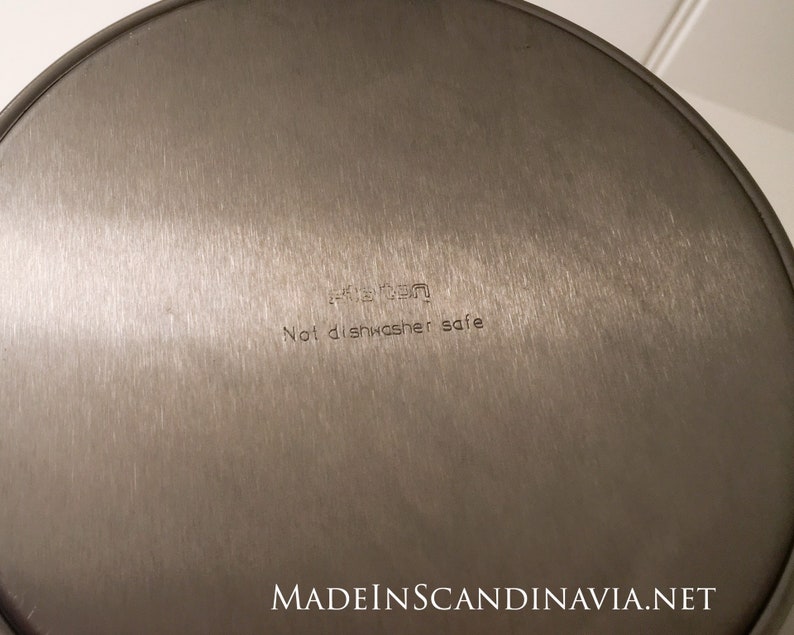 May include: A close-up of the bottom of a stainless steel pan. The text "Not dishwasher safe" is etched into the metal. The pan has a brushed finish and a circular shape. The brand name is also visible.