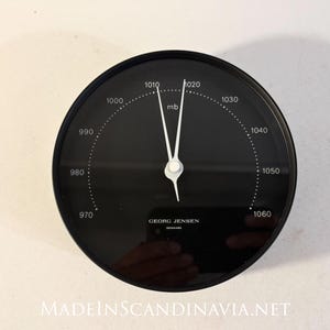 May include: A circular black Georg Jensen barometer with a white scale ranging from 970 to 1060 mb. The device features two thin white hands pointing near the 1010 and 1020 marks, with the brand name Georg Jensen Denmark printed in white text.