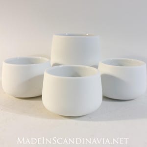 Pode incluir: Quatro xícaras de cerâmica brancas com um design arredondado e minimalista. As xícaras estão dispostas sobre uma superfície branca, com o texto "MADEINSCANDINAVIA.NET" na parte inferior. As xícaras têm um acabamento liso e fosco.