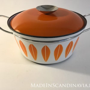 Op de afbeelding: Een oranje en witte geëmailleerde pan met een deksel en zilveren handvatten. De pan heeft een bladmotief.