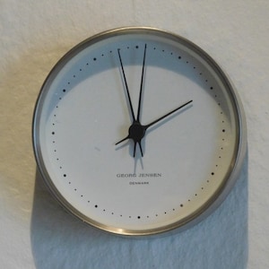 May include: A round, silver-toned wall clock with a white face. The clock features black hands and small black dots marking the hours. The text "GEORG JENSEN DENMARK" is printed at the bottom of the clock face.