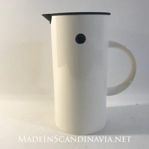 Pode incluir: Uma jarra alta de cerâmica branca com tampa preta e alça arredondada. Um pequeno detalhe circular preto está na lateral. A jarra tem um design minimalista.