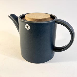 Könnte beinhalten: Eine dunkelblaue Keramik-Teekanne mit zylindrischem Körper, geschwungenem Griff und kurzem Ausguss. Die Teekanne hat einen runden Deckel aus hellem Holz und einen kleinen runden Aufkleber an der Seite mit dem aufgedruckten Text HÖGANÄS KERAMIK.