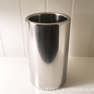 May include: A stainless steel wine chiller with a cylindrical shape. The wine cooler has a polished, reflective surface and a wide opening at the top. The product is designed to keep wine bottles cold.