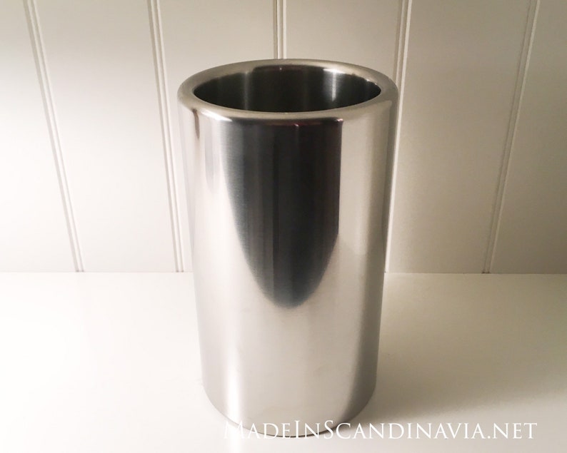 May include: A stainless steel wine chiller with a cylindrical shape. The wine cooler has a polished, reflective surface and a wide opening at the top. The product is designed to keep wine bottles cold.