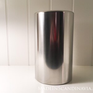 May include: A tall, cylindrical stainless steel ice bucket with a polished finish. The bucket has a rounded base and reflects the surrounding environment. The background is a white surface and wall.
