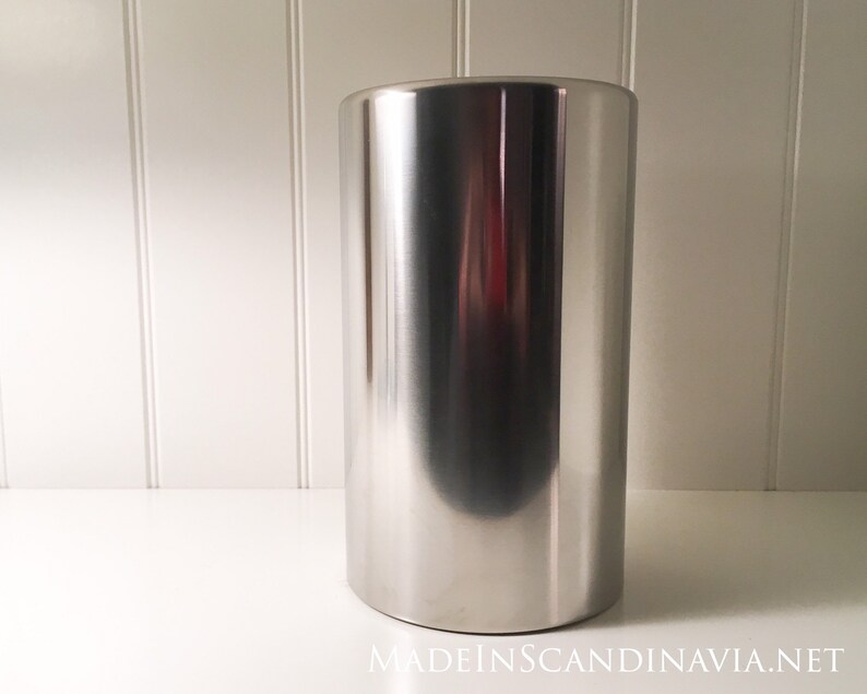 May include: A tall, cylindrical stainless steel ice bucket with a polished finish. The bucket has a rounded base and reflects the surrounding environment. The background is a white surface and wall.