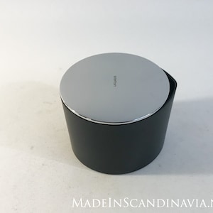 Puede incluir: Un recipiente cilíndrico negro y plateado con tapa. La tapa tiene la palabra "Stelton" impresa.