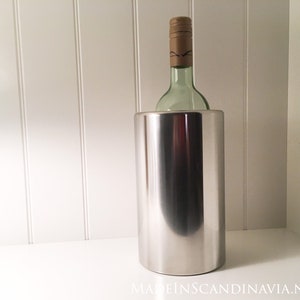 May include: A stainless steel wine chiller with a bottle of wine inside. The wine bottle is green with a brown cap. The chiller is cylindrical and reflects the light. The background is a white wall and shelf.