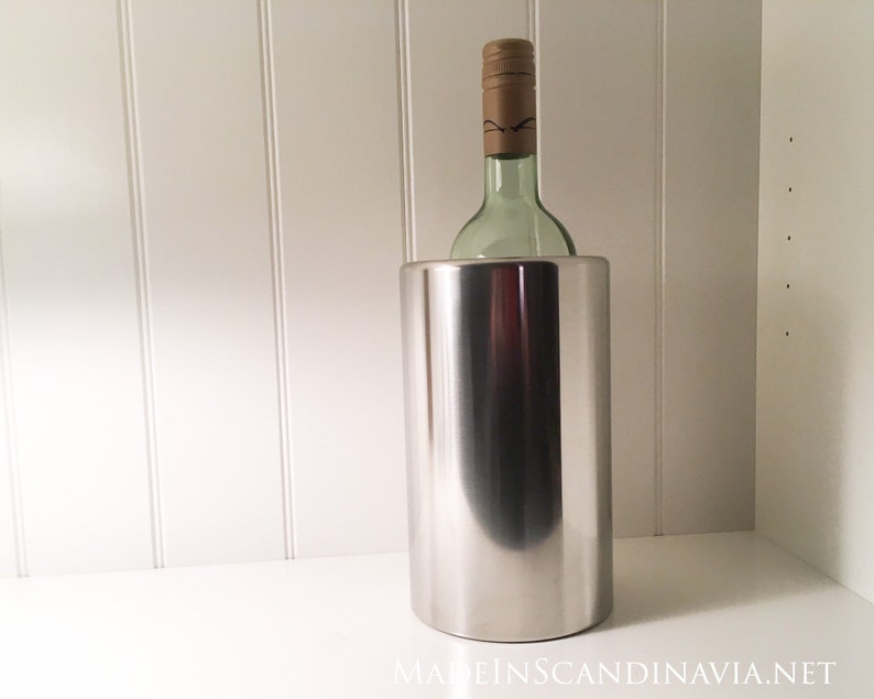 May include: A stainless steel wine chiller with a bottle of wine inside. The wine bottle is green with a brown cap. The chiller is cylindrical and reflects the light. The background is a white wall and shelf.
