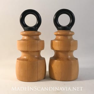 May include: Two wooden door stops with a natural wood finish and black ring handles. The door stops have a turned design and are displayed against a white background. The text "MADEINSCANDINAVIA.NET" is visible at the bottom.
