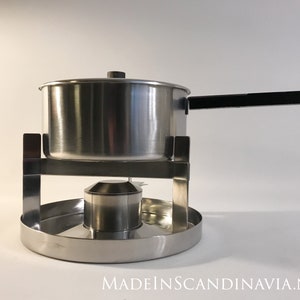 Può includere: Una fonduta in acciaio inossidabile con manico nero e bruciatore. La pentola è appoggiata su una base rotonda in acciaio inossidabile.