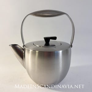 May include: A stainless steel kettle with a curved handle and a black lid handle. The kettle has a rounded body and a spout for pouring. The text "MADEINSCANDINAVIA.NET" is at the bottom of the image.