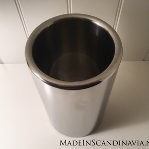 May include: A stainless steel wine chiller with a polished finish. The cylindrical container has a wide opening and a slightly tapered body. The text "MADEINSCANDINAVIA.NET" is visible at the bottom.