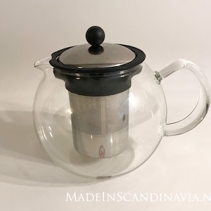 May include: A clear glass teapot with a black lid and a stainless steel infuser. The teapot has a round body and a curved handle.