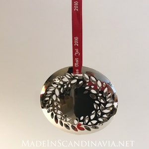 Könnte beinhalten: Silberfarbener Weihnachtsschmuck in Kreisform mit ausgeschnittenem Lorbeerkranz-Design. Er hängt an einem roten Band mit der Aufschrift "Noël Jul 2016". Am unteren Rand steht der Text "MADEINSCANDINAVIA.NET".