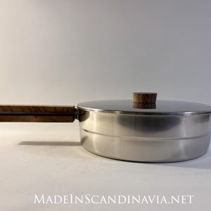 Puede incluir: Una cacerola de acero inoxidable con tapa y asa de madera. El asa es de color marrón oscuro y está sujeta al lateral de la cacerola. La tapa tiene un pomo de madera. El texto "MADEINSCANDINAVIA.NET" está en la parte inferior de la imagen.