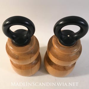 May include: Two wooden salt and pepper shakers with a light wood finish and black handles. The shakers have a stacked, rounded design and are displayed on a white surface. The text "MADEINSCANDINAVIA.NET" is at the bottom.