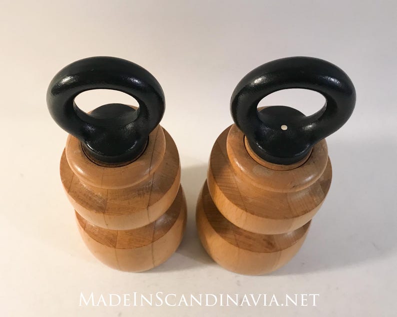 May include: Two wooden salt and pepper shakers with a light wood finish and black handles. The shakers have a stacked, rounded design and are displayed on a white surface. The text "MADEINSCANDINAVIA.NET" is at the bottom.
