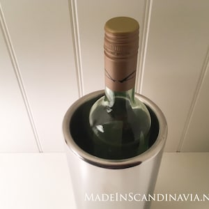 May include: A stainless steel wine chiller holds a green glass wine bottle with a brown and gold cap. The chiller is cylindrical and reflects the light. The bottle is partially visible, suggesting a chilled beverage.