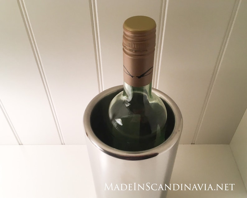 May include: A stainless steel wine chiller holds a green glass wine bottle with a brown and gold cap. The chiller is cylindrical and reflects the light. The bottle is partially visible, suggesting a chilled beverage.